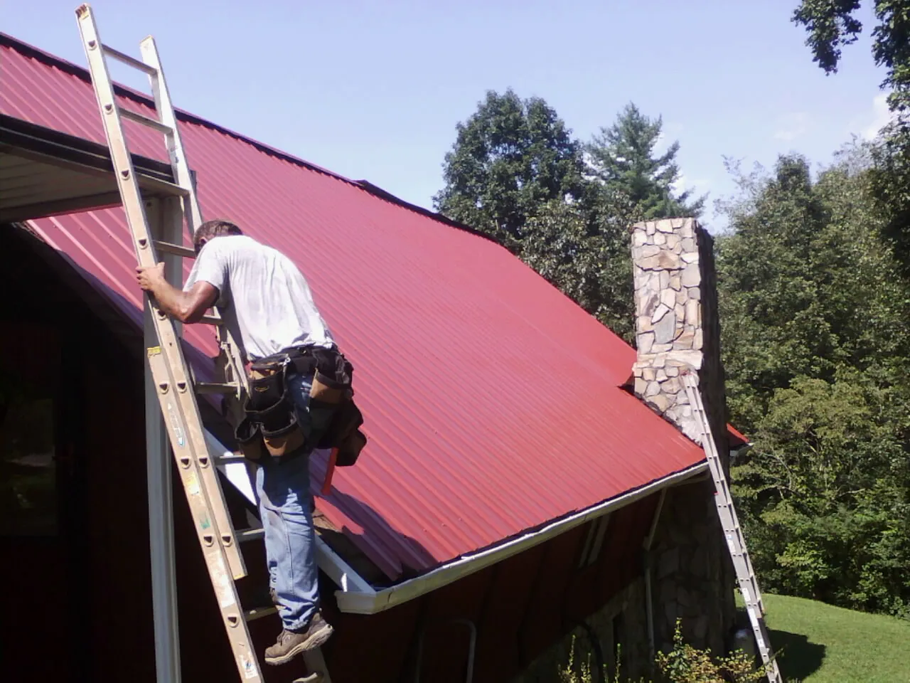 Roofer on a ladder beside a red metal roof and stone chimney — Soffit & Fascia Repair by Roofing Brothers in Marsing